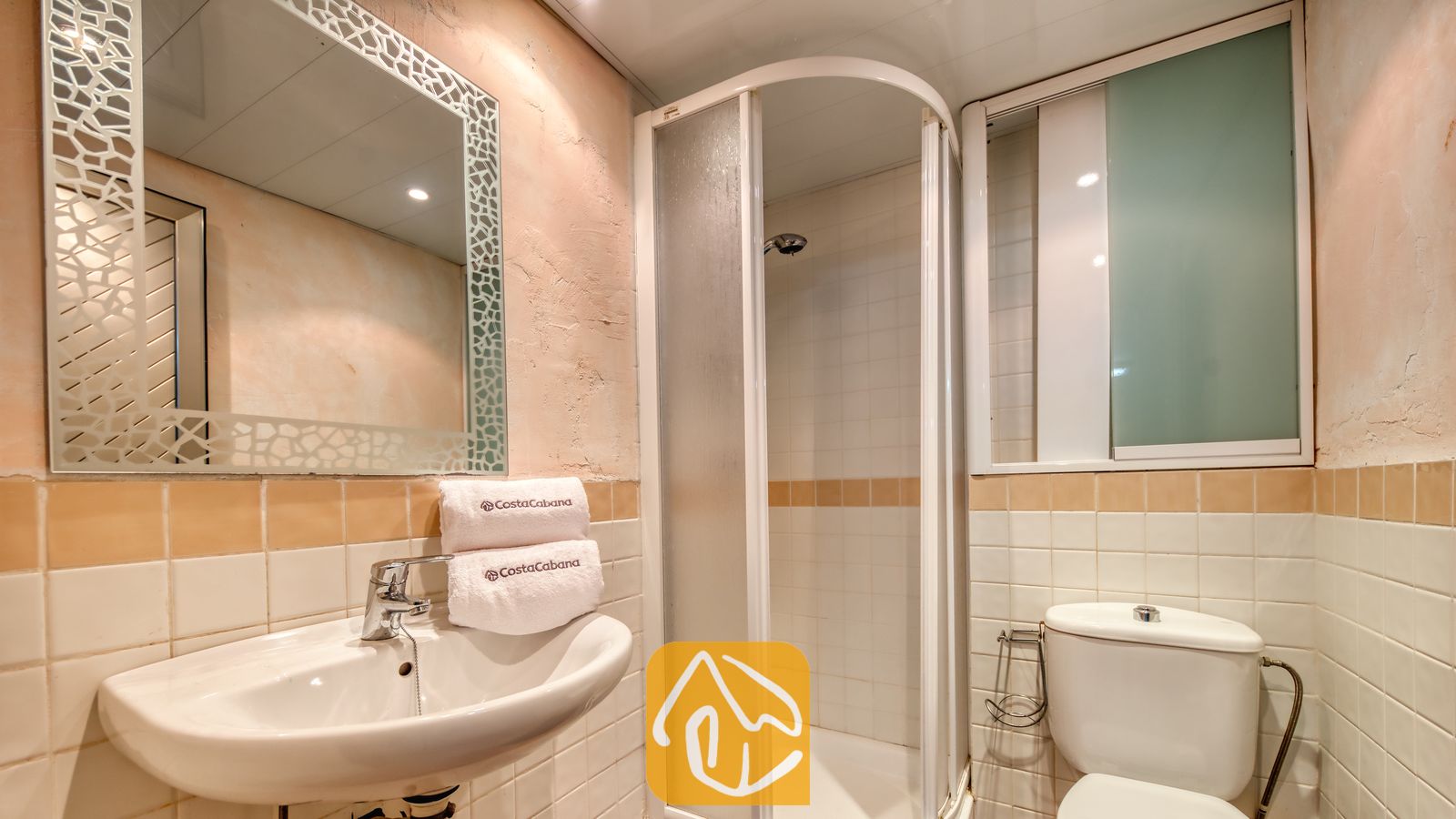 Vakantievilla's Costa Brava Spanje - Villa Sandy Beach - Badkamer van Villa Sandy Beach met een witte wastafel op zuil, een hoekdouche en een grote spiegel met een decoratieve, gematteerde rand. De onderste helft van de muren is betegeld met crèmekleurige tegels, afgewerkt met een zandkleurige sierstrip.