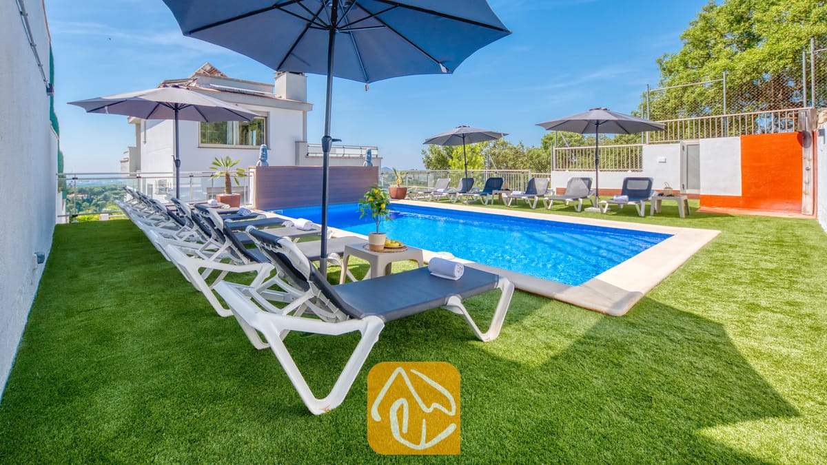 Vakantievilla's Costa Brava Spanje - Villa Sandy Beach - Privézwembad bij Villa Sandy Beach met rijen witte ligbedden, blauwe parasols en een groen kunstgrasveld onder een strakblauwe lucht.