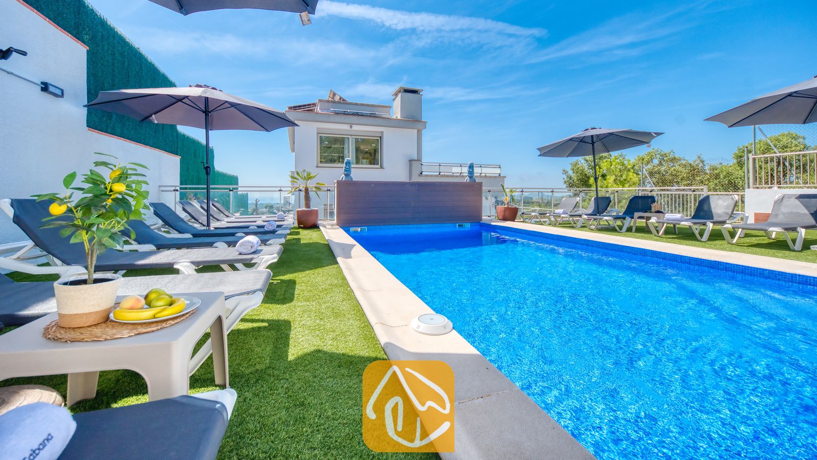 Vakantievilla's Costa Brava Spanje - Villa Sandy Beach - Privézwembad bij Villa Sandy Beach, omringd door kunstgras, grijze ligbedden met witte handdoeken en donkere parasols onder een strakblauwe lucht.