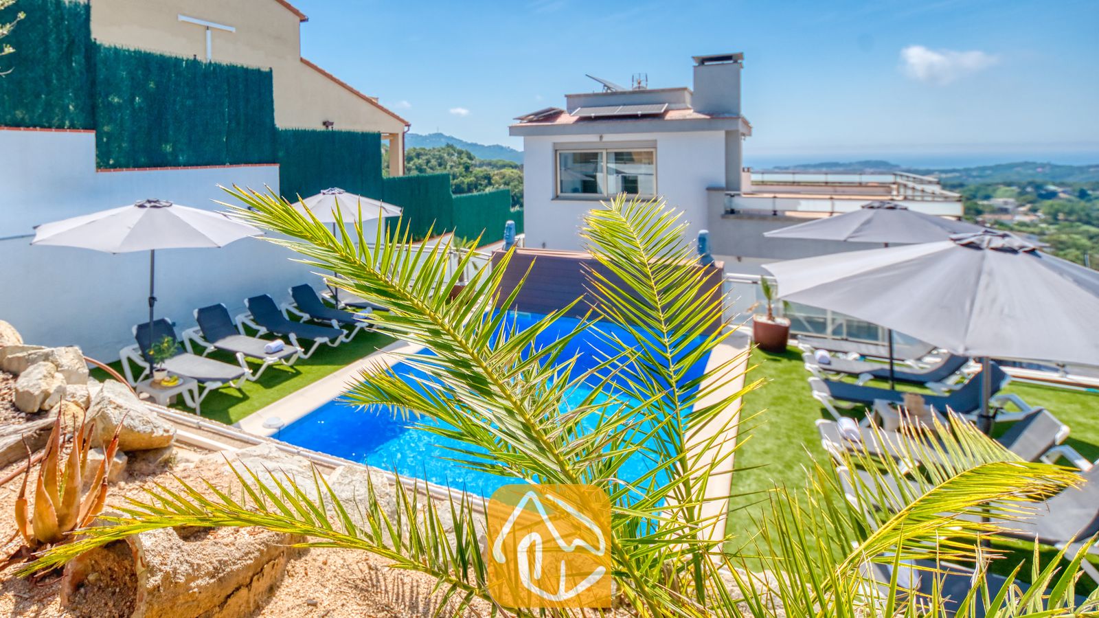 Vakantievilla's Costa Brava Spanje - Villa Sandy Beach - Buitenzwembad bij Villa Sandy Beach met grijze ligbedden op een groen gazon, witte parasols en een weids uitzicht op de kust onder een strakblauwe lucht.