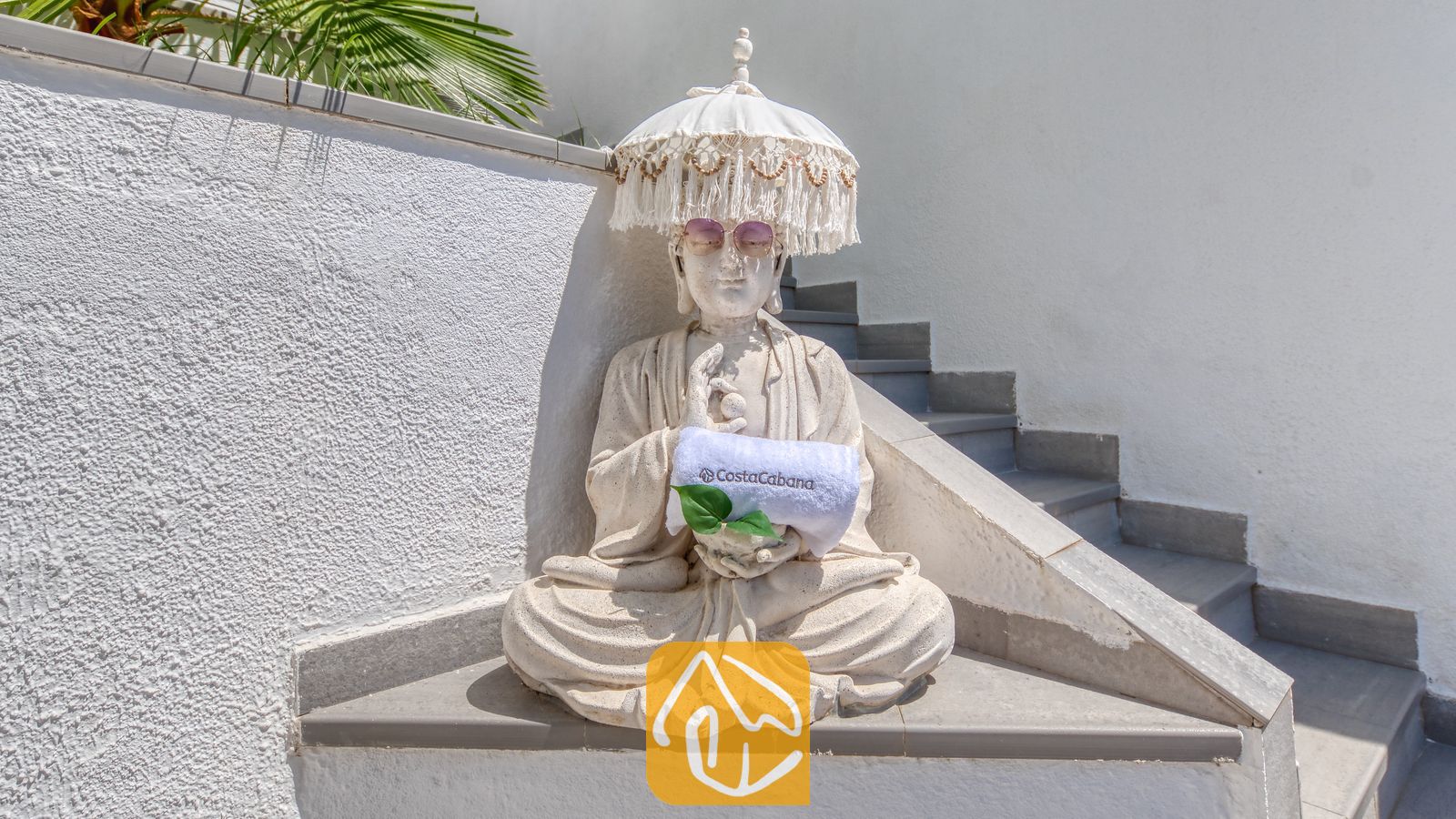Vakantievilla's Costa Brava Spanje - Villa Sandy Beach - Stenen Boeddhabeeld met zonnebril en een witte parasolhoed met franjes, dat een opgerolde handdoek vasthoudt naast de buitentrap van Villa Sandy Beach.