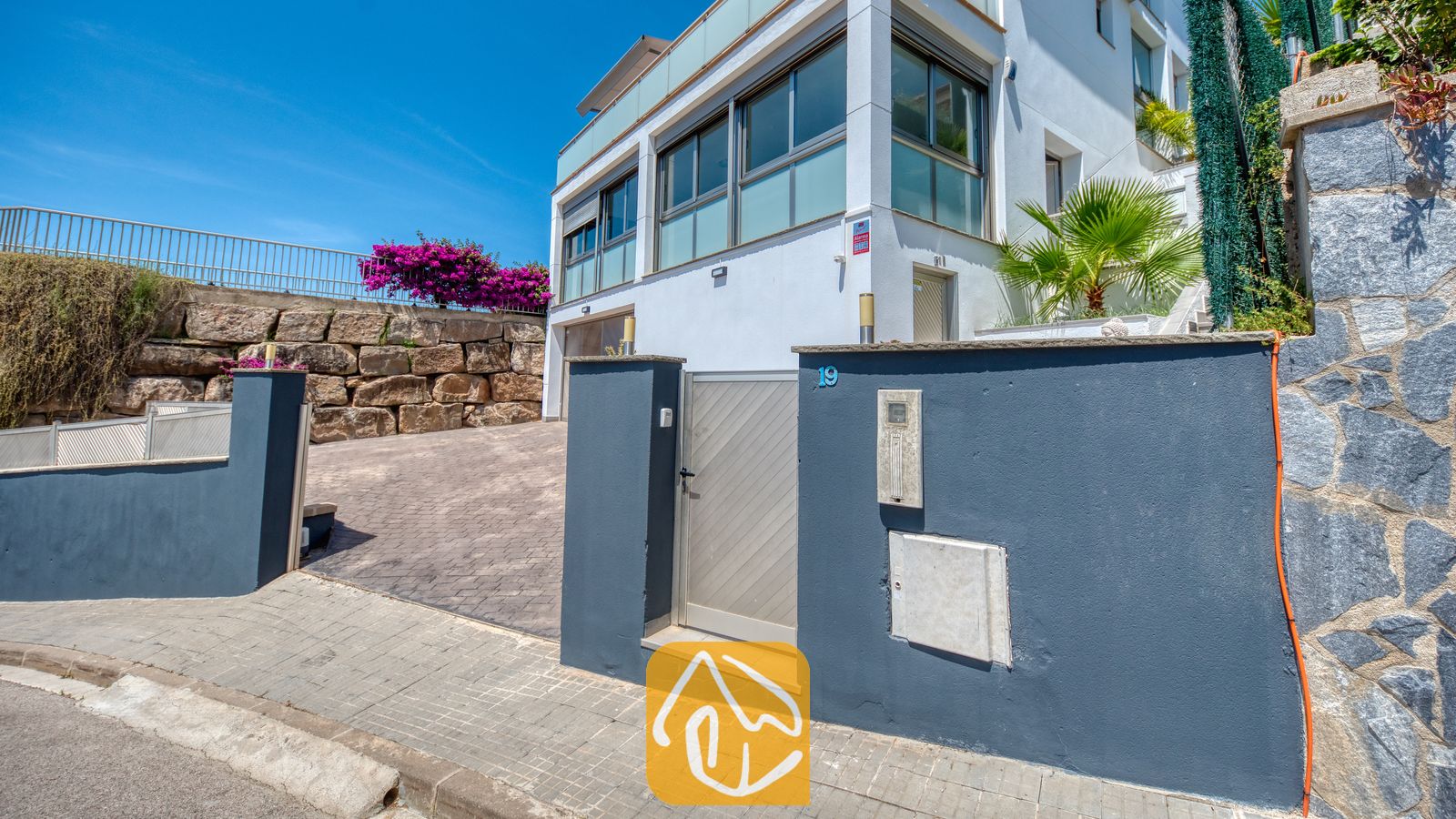 Vakantievilla's Costa Brava Spanje - Villa Sandy Beach - De omheinde entree van Villa Sandy Beach beschikt over een grijze muur, een zilverkleurig hek en een verharde oprit die leidt naar een wit gebouw van meerdere verdiepingen met grote raampartijen en een stenen keermuur.