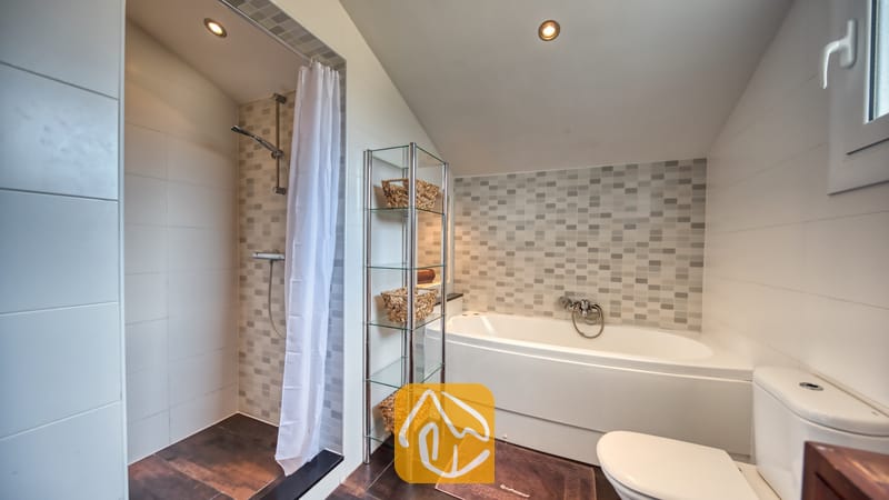 Villas de vacances Costa Brava Espagne - Villa Madonna - Salle de bains moderne comprenant une douche à l'italienne carrelée avec rideau blanc, une baignoire blanche, une étagère en verre avec des paniers tressés et des toilettes.