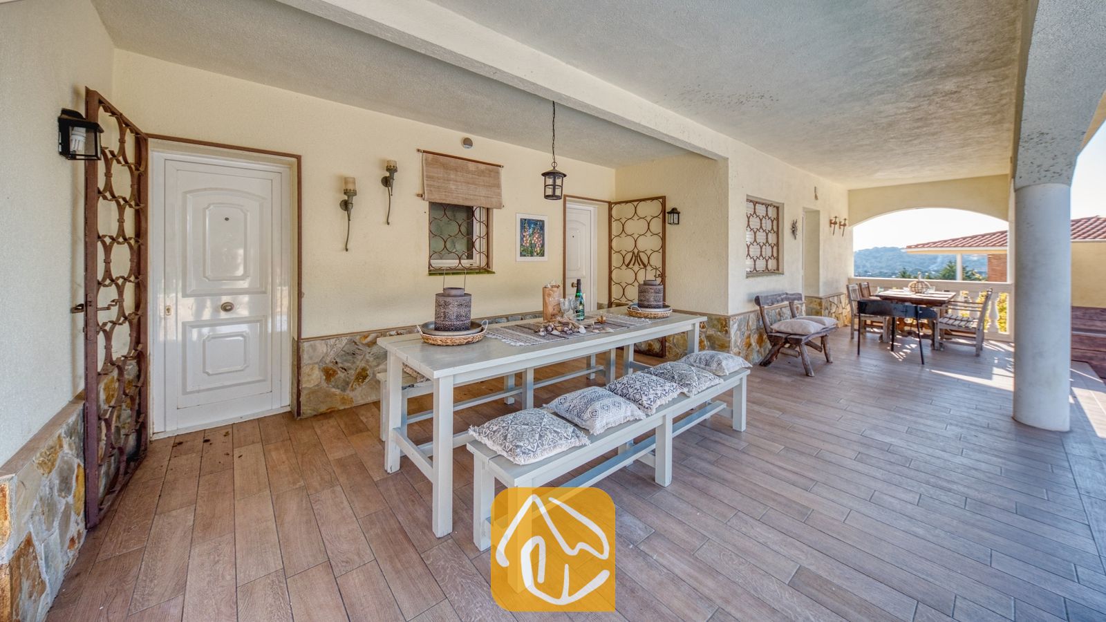 Vakantievilla's Costa Brava Spanje - Villa Madonna - Overdekt buitenterras bij Villa Madonna met een lange witte eettafel met bijpassende banken, kussens met een patroon, stenen funderingsmuren en een tegelvloer.