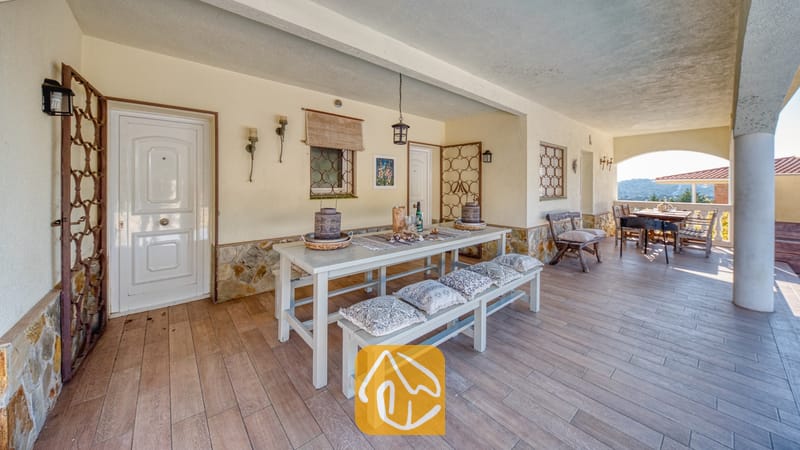 Vakantievilla's Costa Brava Spanje - Villa Madonna - Overdekt buitenterras bij Villa Madonna met een lange witte eettafel met bijpassende banken, kussens met een patroon, stenen funderingsmuren en een tegelvloer.