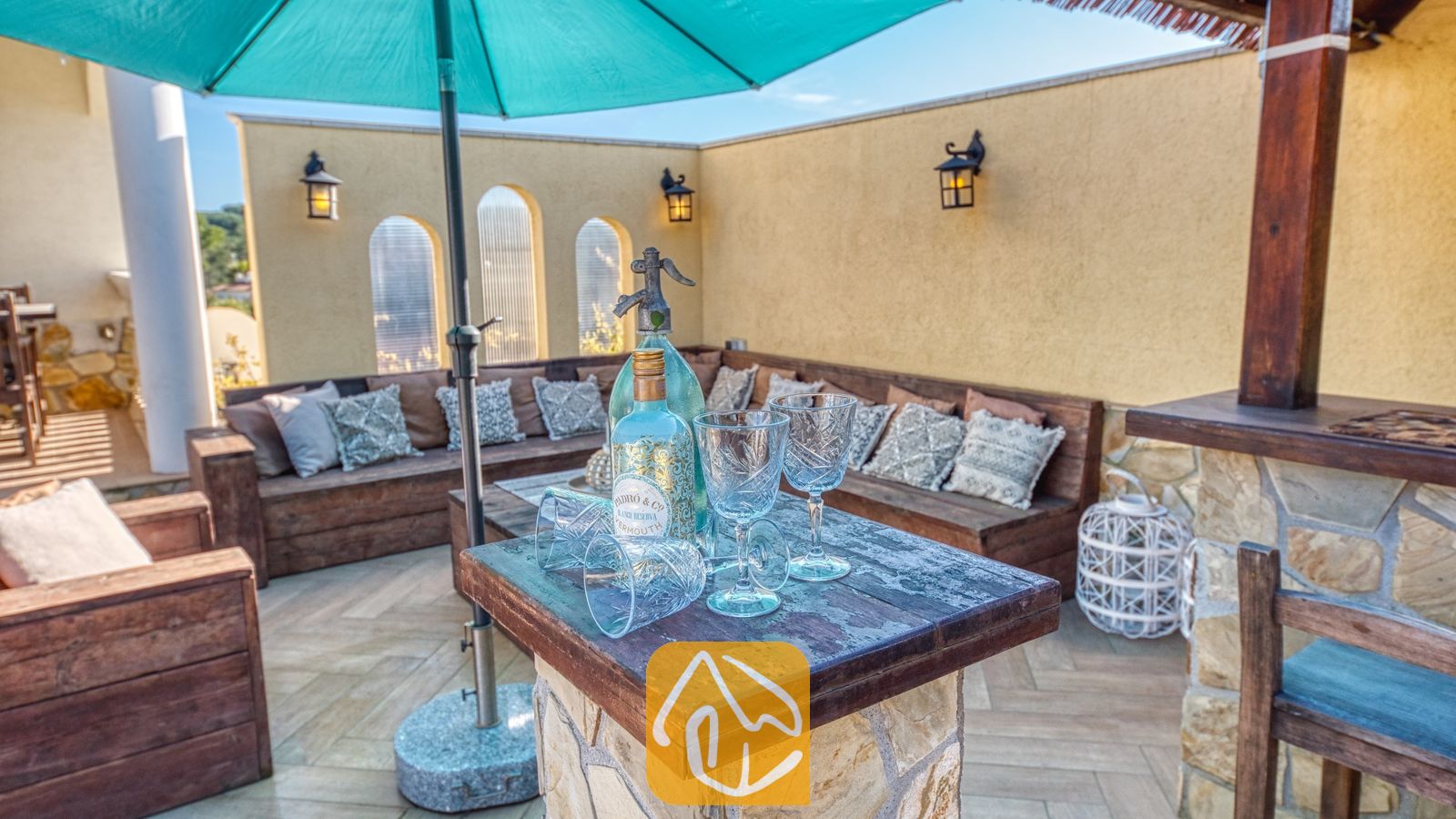 Vakantievilla's Costa Brava Spanje - Villa Madonna - Buitenbar bij Villa Madonna met een stenen barblad, glaswerk, flessen, een grote blauwgroene parasol en houten banken met gedessineerde kussens.