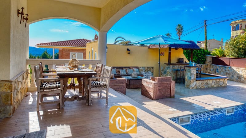 Vakantievilla's Costa Brava Spanje - Villa Madonna - Overdekt terras bij Villa Madonna met een rustieke houten eettafel, fauteuils en een betegeld buitenzwembad met een blauwe parasol.