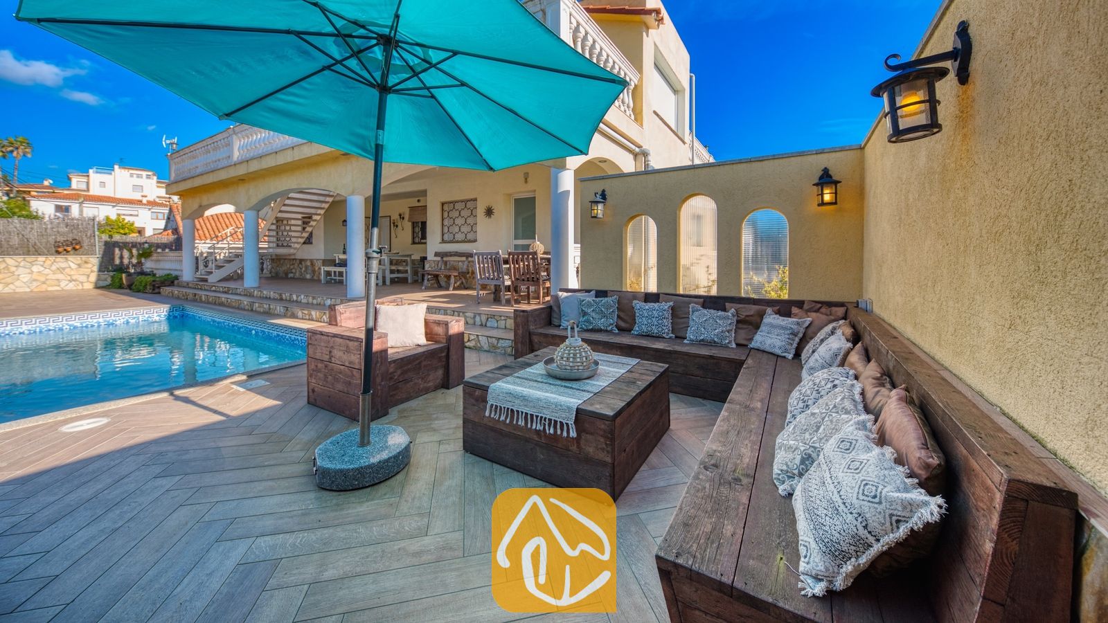 Vakantievilla's Costa Brava Spanje - Villa Madonna - Houten buitenloungehoek met bedrukte kussens en een grote blauwe parasol bij het zwembad van Villa Madonna.