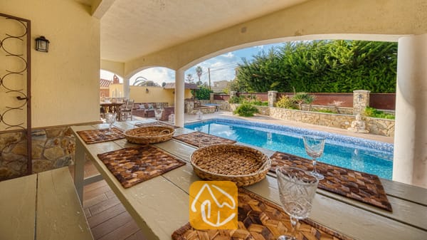 Vakantievilla's Costa Brava Spanje - Villa Madonna - Overdekte eettafel buiten met gevlochten placemats en wijnglazen bij Villa Madonna, met uitzicht op een blauw betegeld zwembad en tuinterras.