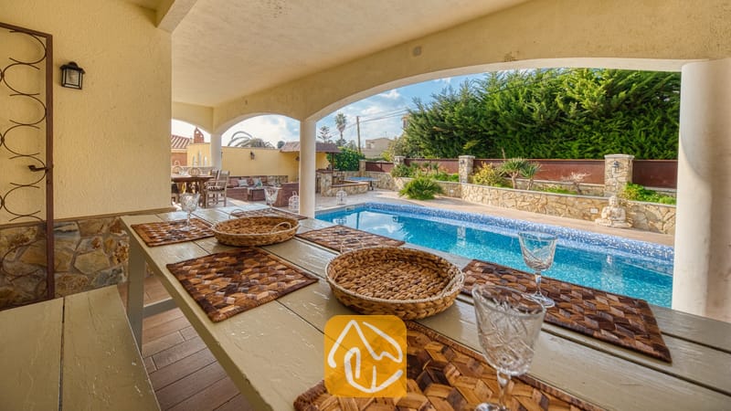 Vakantievilla's Costa Brava Spanje - Villa Madonna - Overdekte eettafel buiten met gevlochten placemats en wijnglazen bij Villa Madonna, met uitzicht op een blauw betegeld zwembad en tuinterras.