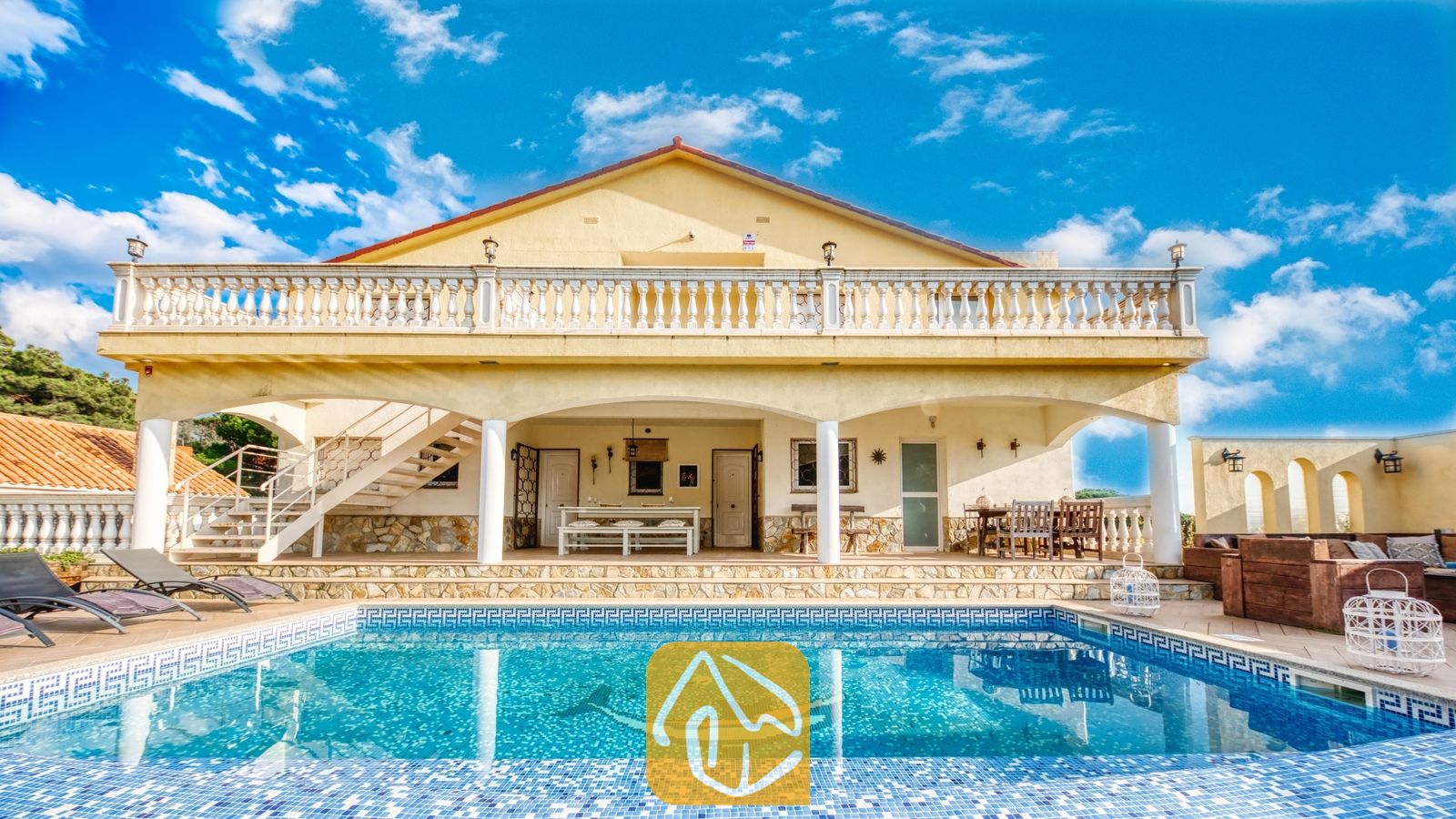 Vakantievilla's Costa Brava Spanje - Villa Madonna - Buitenaanzicht van Villa Madonna met een zwembad voorzien van een dolfijnen-mozaïek tegel, een overdekte veranda, een met steen bekleed terras en gele buitenmuren onder een blauwe lucht.