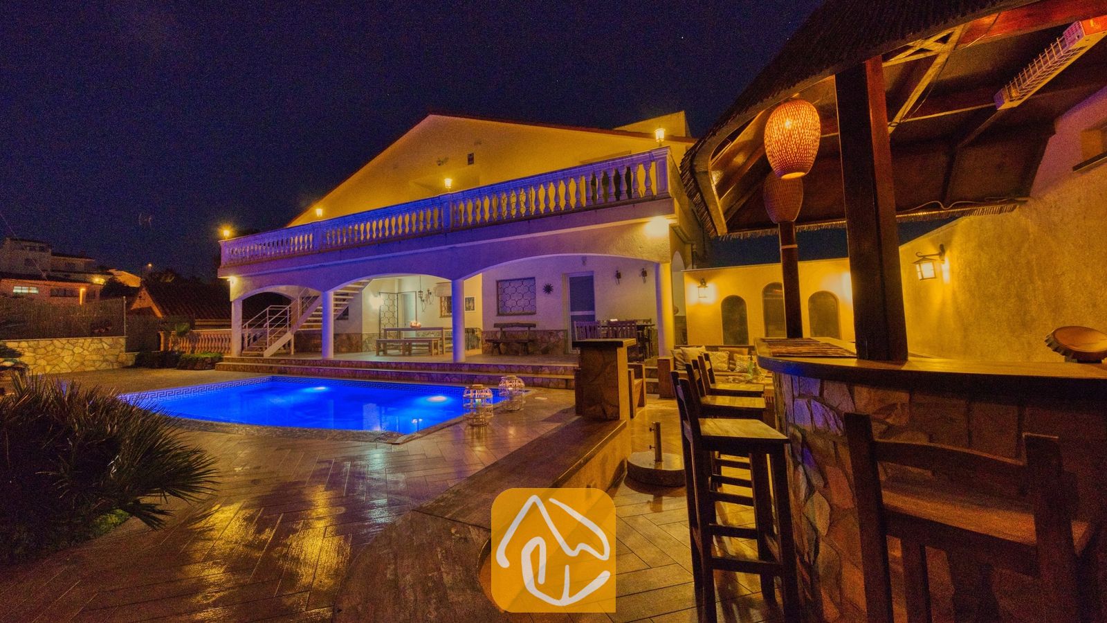 Vakantievilla's Costa Brava Spanje - Villa Madonna - De buitenzijde van Villa Madonna bij nacht, met een verlicht zwembad, een overdekte buitenbar met houten krukken en een verlicht terras met een balustrade.