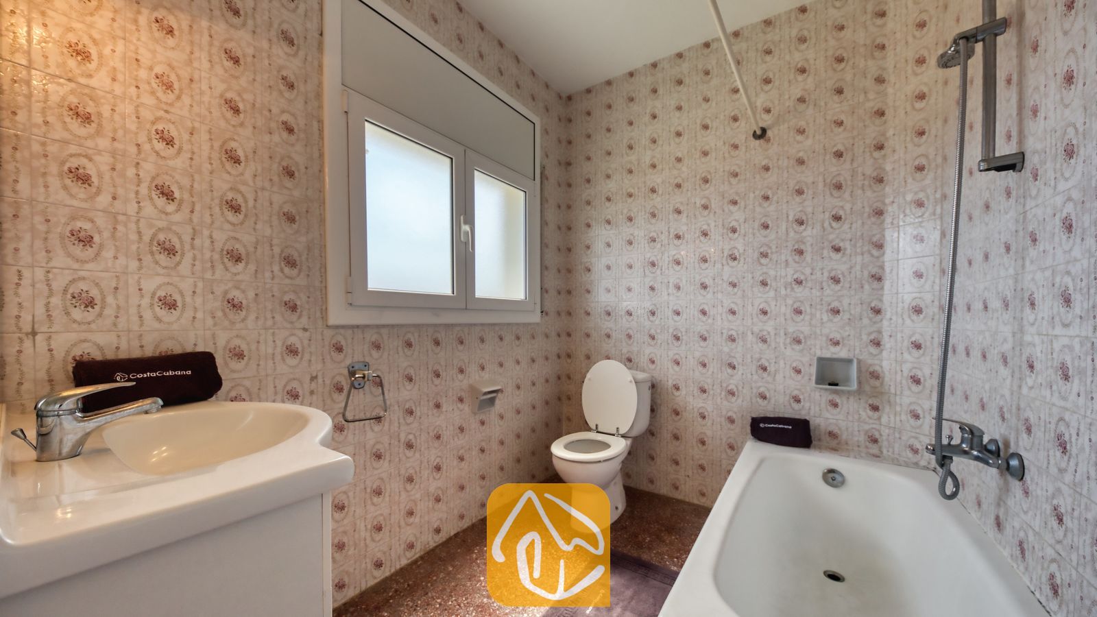 Vakantievilla's Costa Brava Spanje - Villa Madonna - Badkamer voorzien van muren met patroon van keramische tegels, een witte wastafel, een ligbad met een verticale douchekop en een toilet onder een matglas raam.