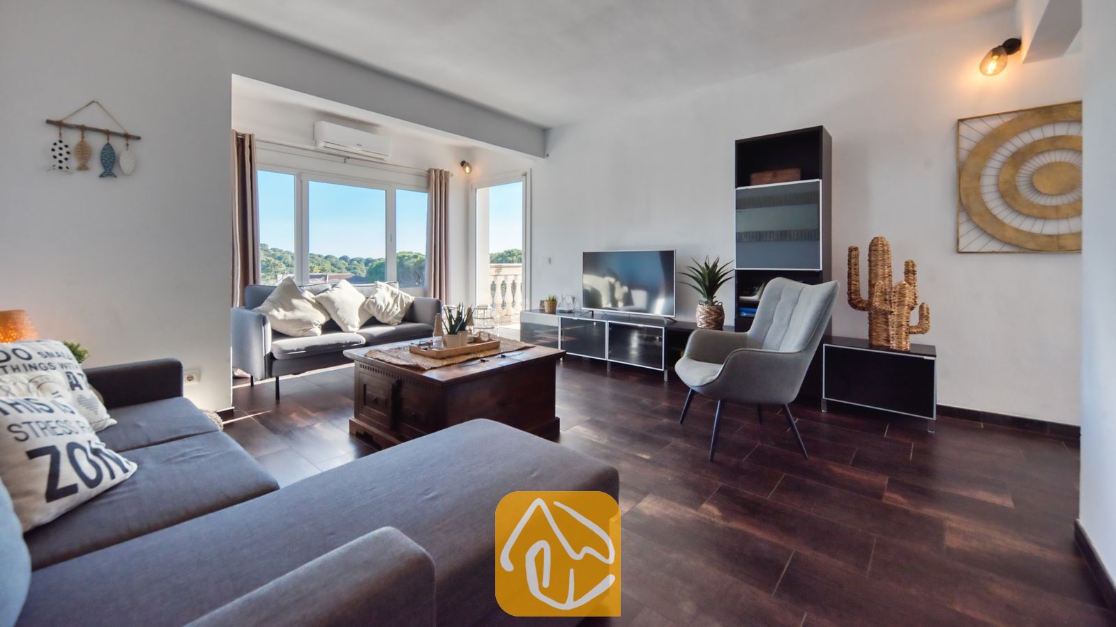 Vakantievilla's Costa Brava Spanje - Villa Madonna - Woonkamer van Villa Madonna met donkere houten vloeren, een grijze hoekbank, een houten salontafel en grote ramen met uitzicht op het groen.