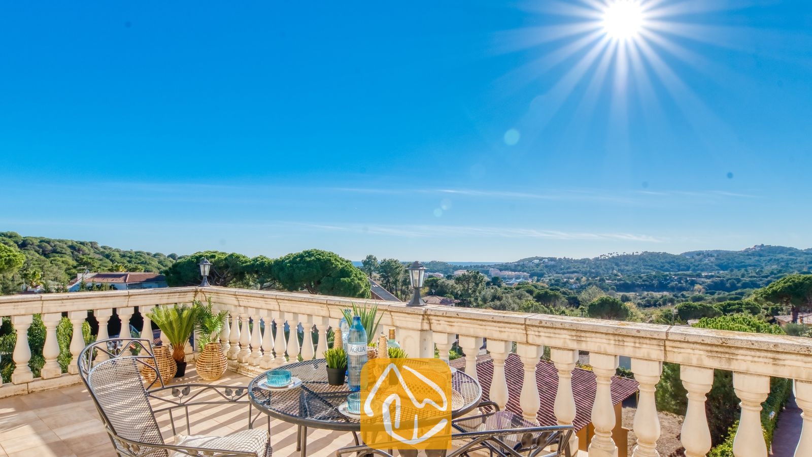Vakantievilla's Costa Brava Spanje - Villa Madonna - Zonnig terras bij Villa Madonna met een smeedijzeren tafel, strooien hoed en een prachtig uitzicht over de groene vallei onder een helderblauwe hemel.