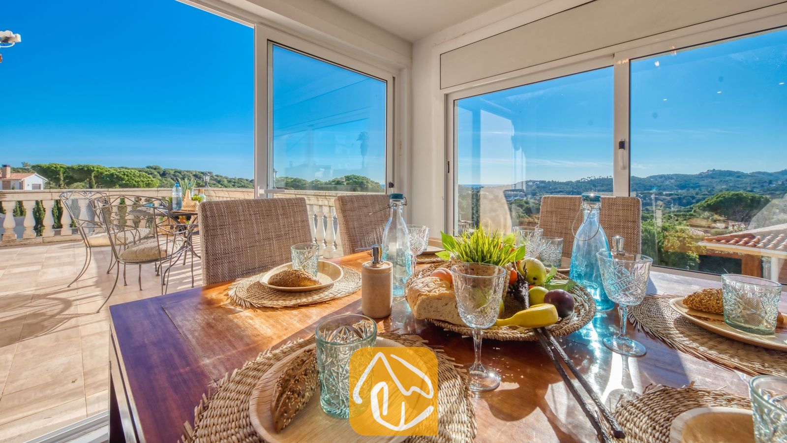 Vakantievilla's Costa Brava Spanje - Villa Madonna - Eettafel binnen in Villa Madonna, gedekt met gevlochten placemats, glaswerk, brood en fruit, omringd door grote glazen ramen met uitzicht op een terras en een groen landschap.