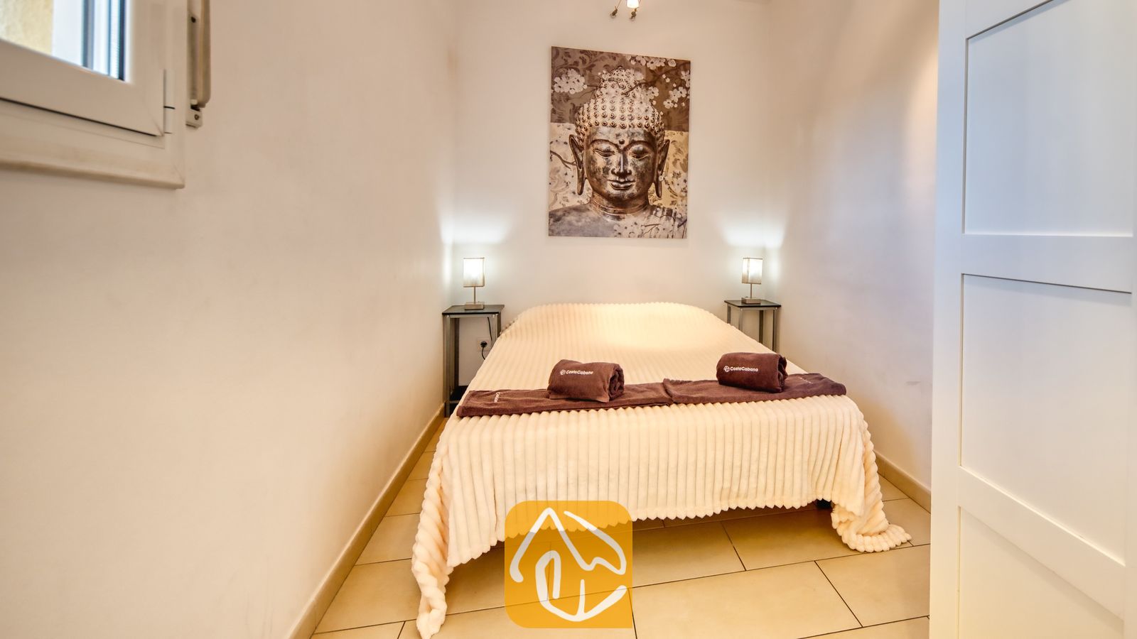 Vakantievilla's Costa Brava Spanje - Villa Madonna - Slaapkamer met een tweepersoonsbed voorzien van een lichtkleurige, geribbelde sprei, opgerolde bruine handdoeken en een groot wandkleed met een Boeddha-afbeelding erboven.