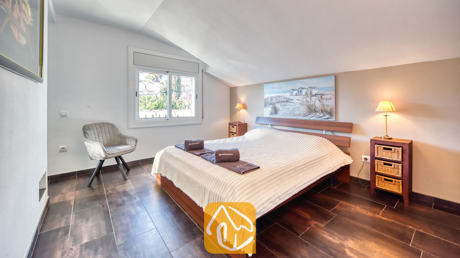 Vakantievilla's Costa Brava Spanje - Villa Madonna - Slaapkamer met een houten bedframe, een wit structuurdekbed, twee nachtkastjes met rieten lades en een grijze fauteuil bij het raam.