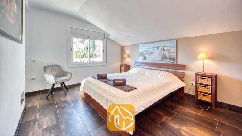 Villas de vacances Costa Brava Espagne - Villa Madonna - Chambre à coucher dotée d'un cadre de lit en bois, d'une couette blanche texturée, de deux tables de chevet avec des tiroirs en osier surmontées de lampes, et d'un fauteuil gris placé près d'une fenêtre.