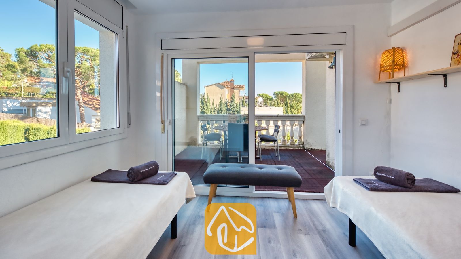 Vakantievilla's Costa Brava Spanje - Villa Madonna - Tweepersoonskamer met twee aparte bedden in Villa Madonna, voorzien van donkergrijze opgerolde handdoeken, een houten bankje en schuifpui naar een balkon.