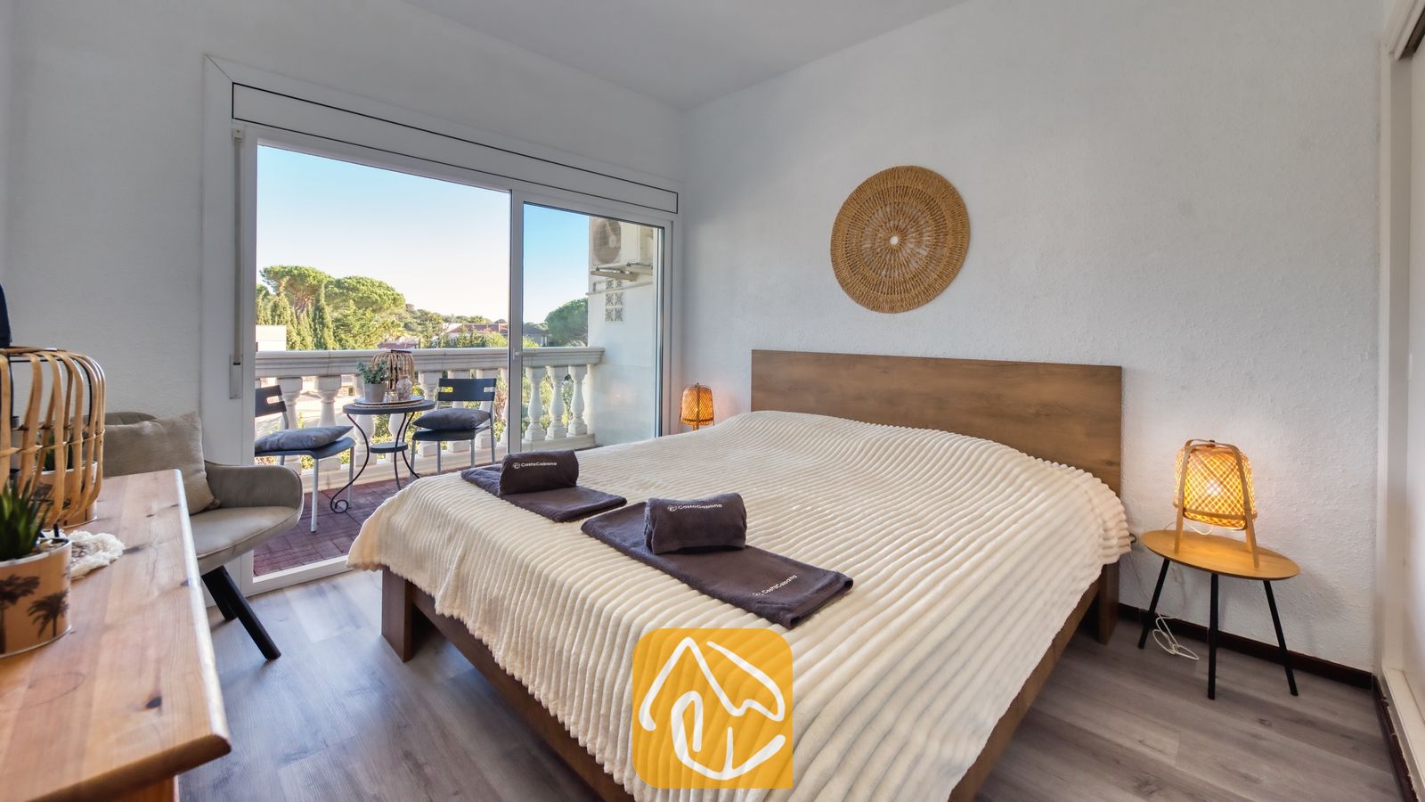 Vakantievilla's Costa Brava Spanje - Villa Madonna - Slaapkamer in Villa Madonna met een houten bed voorzien van een crèmekleurige, geribbelde sprei, geweven bedlampjes en een schuifpui die uitkomt op een balkon.