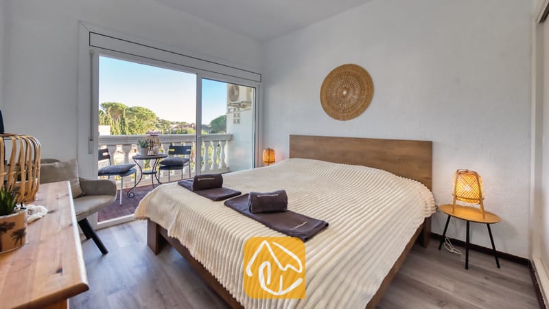 Villas de vacances Costa Brava Espagne - Villa Madonna - Chambre à la Villa Madonna dotée d'un lit en bois avec un plaid côtelé crème, de lampes de chevet tressées et de baies vitrées coulissantes s'ouvrant sur un balcon.