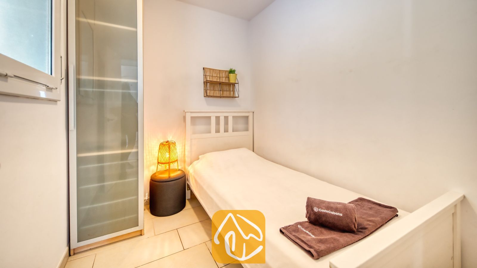 Vakantievilla's Costa Brava Spanje - Villa Madonna - Slaapkamer met een wit houten bed, bruine opgevouwen handdoeken, een geweven bedlampje op een donkere kruk en een hoge kledingkast van matglas.