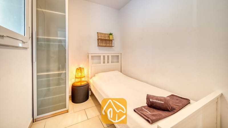 Villas de vacances Costa Brava Espagne - Villa Madonna - Chambre simple dotée d'un lit en bois blanc, de serviettes pliées marron, d'une lampe de chevet tressée posée sur un tabouret foncé et d'une grande armoire en verre dépoli.