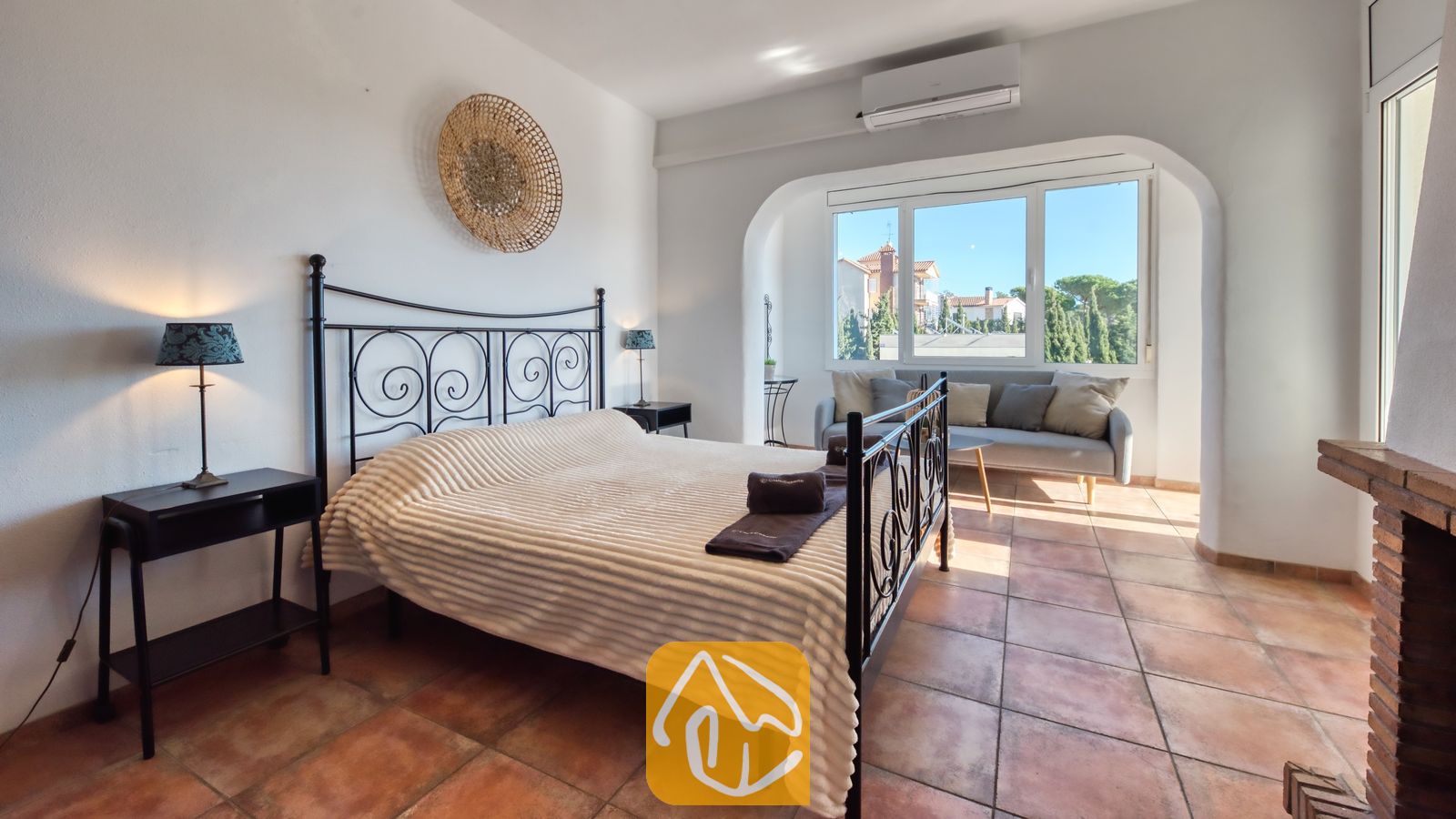 Vakantievilla's Costa Brava Spanje - Villa Madonna - Slaapkamer met een zwart metalen bedframe, een wit geweven deken en terracotta plavuizen.