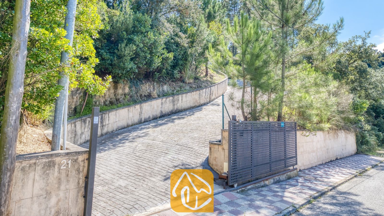 Vakantievilla's Costa Brava Spanje - Villa Esmee - De hellende, verharde oprit van Villa Esmee leidt omhoog door een stenen keermuur en omringende beplanting, voorzien van een metalen toegangspoort.
