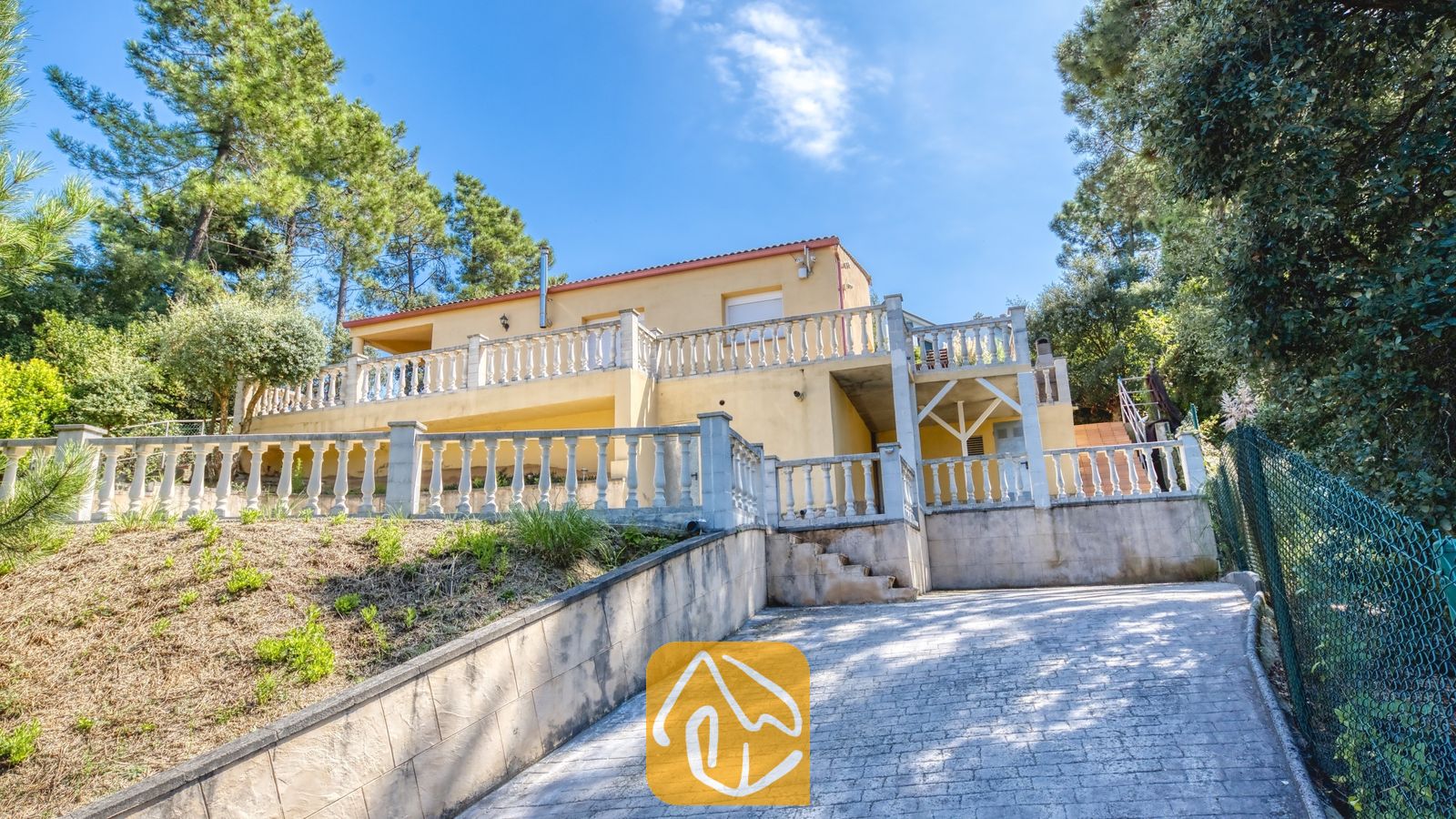 Vakantievilla's Costa Brava Spanje - Villa Esmee - Buitengevel van Villa Esmee met een tweedelig geel huis met een stenen balustradebalkon, een bestrate oprijlaan en omringende dennenbomen onder een blauwe hemel.