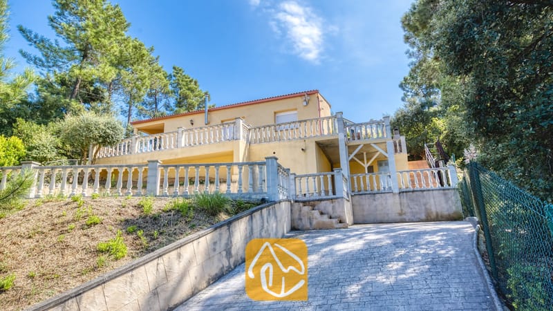 Villas de vacances Costa Brava Espagne - Villa Esmee - Façade extérieure de la Villa Esmee présentant une maison jaune à deux étages dotée d’un balcon avec balustrade en béton, d’une allée pavée et de pins environnants sous un ciel bleu.