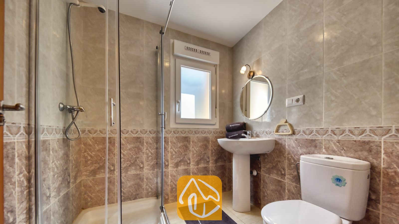 Vakantievilla's Costa Brava Spanje - Villa Esmee - Moderne betegelde badkamer met een glazen inloopdouche, een wastafel met zuil en een ronde spiegel, en een wit toilet tegen beige muren met marmerlook.