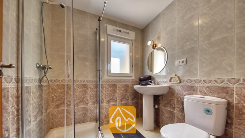 Villas de vacances Costa Brava Espagne - Villa Esmee - Salle de bains moderne carrelée comprenant une douche fermée par une paroi en verre, un lavabo sur pied surmonté d'un miroir rond, et des toilettes blanches sur fond de murs effet marbre beige.