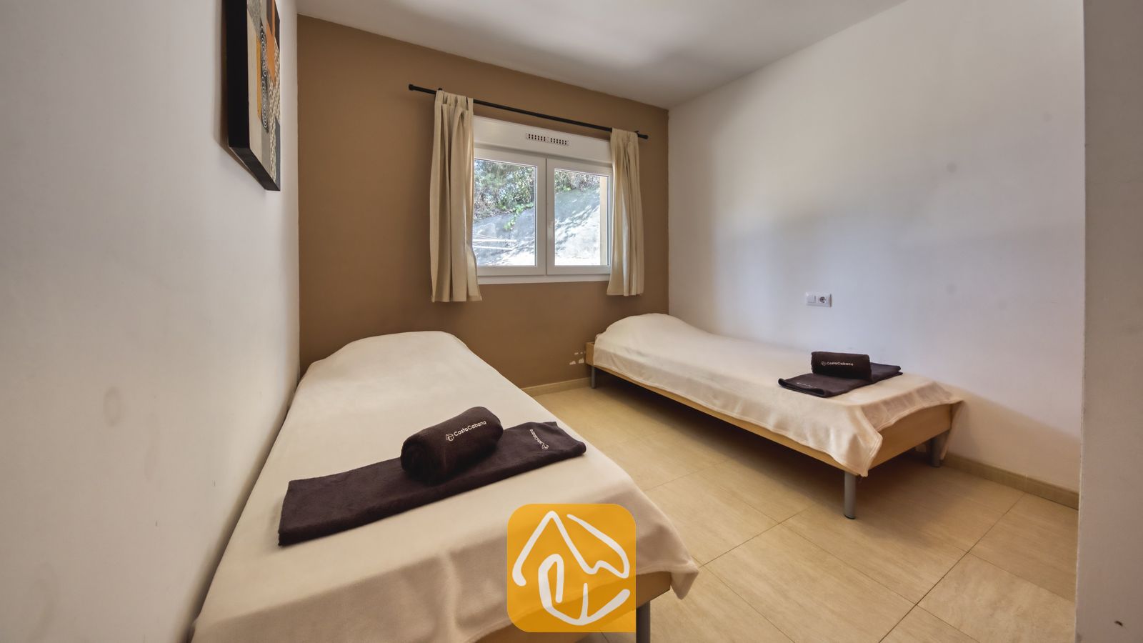 Vakantievilla's Costa Brava Spanje - Villa Esmee - Slaapkamer met twee eenpersoonsbedden voorzien van witte spreien en donkere, opgerolde handdoeken, een raam met beige gordijnen en een accentmuur.