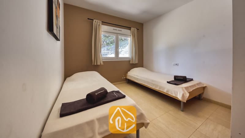 Villas de vacances Costa Brava Espagne - Villa Esmee - Chambre avec deux lits simples habillés de housses blanches et de serviettes roulées sombres, une fenêtre aux rideaux beige et un mur d'accent.
