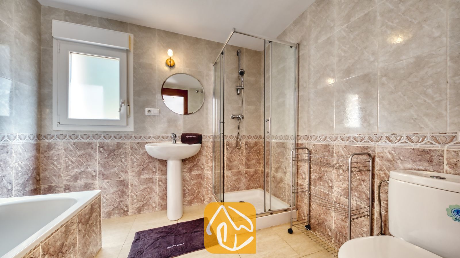 Vakantievilla's Costa Brava Spanje - Villa Esmee - Badkamer voorzien van beige wandtegels met marmerlook, een glazen inloopdouche, een witte wastafel met zuil en ronde spiegel, en een toilet.