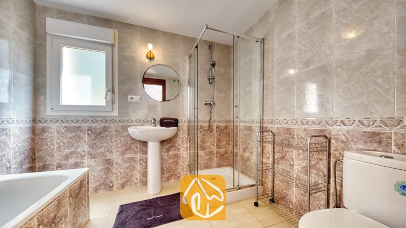 Villas de vacances Costa Brava Espagne - Villa Esmee - Salle de bains comprenant des carreaux muraux beiges effet marbre, une douche d'angle vitrée, un lavabo blanc sur pied surmonté d'un miroir rond, ainsi qu'une toilettes.
