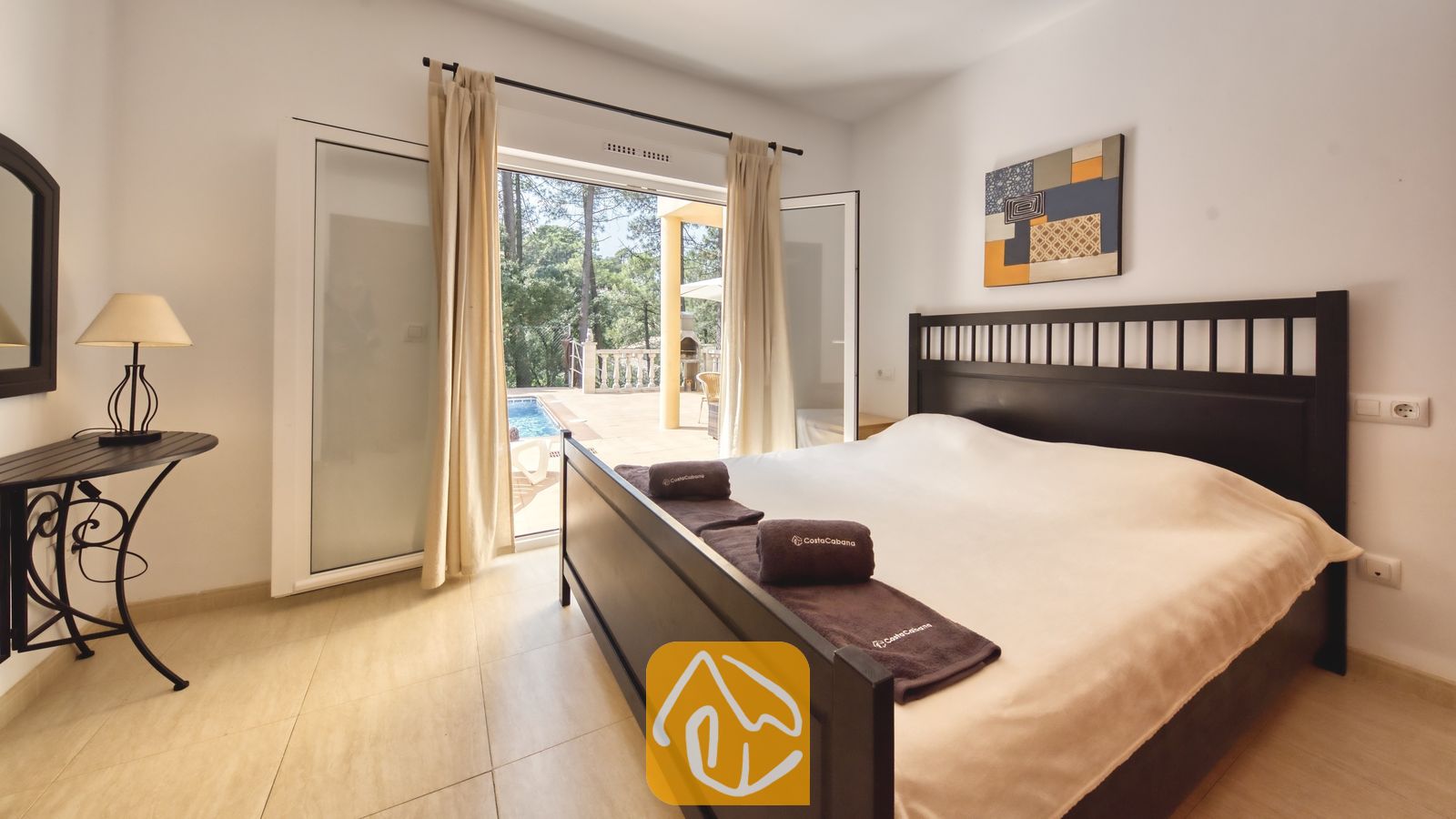 Vakantievilla's Costa Brava Spanje - Villa Esmee - Slaapkamer met een donker houten bedframe, opgevouwen bruine handdoeken en openslaande glazen deuren naar een zonnig terras met een zwembad.