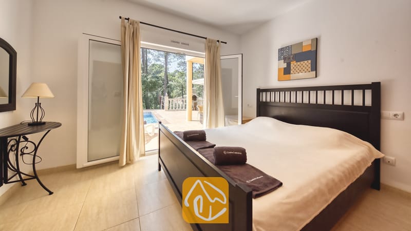 Vakantievilla's Costa Brava Spanje - Villa Esmee - Slaapkamer met een donker houten bedframe, opgevouwen bruine handdoeken en openslaande glazen deuren naar een zonnig terras met een zwembad.
