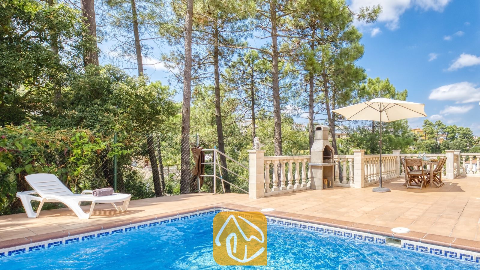 Vakantievilla's Costa Brava Spanje - Villa Esmee - Buitenterras van Villa Esmee met een betegelde zwembadrand, witte ligstoel, stenen barbecue en een eettafel onder een parasol tegen een achtergrond van dennenbomen.