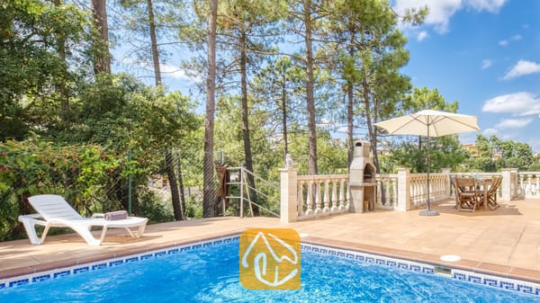 Vakantievilla's Costa Brava Spanje - Villa Esmee - Buitenterras van Villa Esmee met een betegelde zwembadrand, witte ligstoel, stenen barbecue en een eettafel onder een parasol tegen een achtergrond van dennenbomen.
