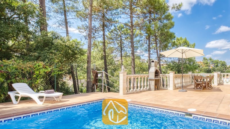 Vakantievilla's Costa Brava Spanje - Villa Esmee - Buitenterras van Villa Esmee met een betegelde zwembadrand, witte ligstoel, stenen barbecue en een eettafel onder een parasol tegen een achtergrond van dennenbomen.