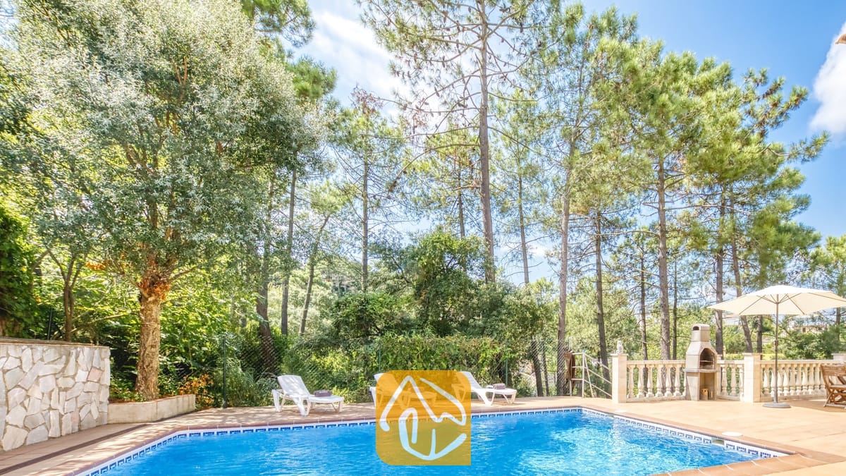 Vakantievilla's Costa Brava Spanje - Villa Esmee - Privézwembad bij Villa Esmee met witte ligstoelen op het betegelde terras, omringd door hoge dennenbomen en een stenen barbecue.