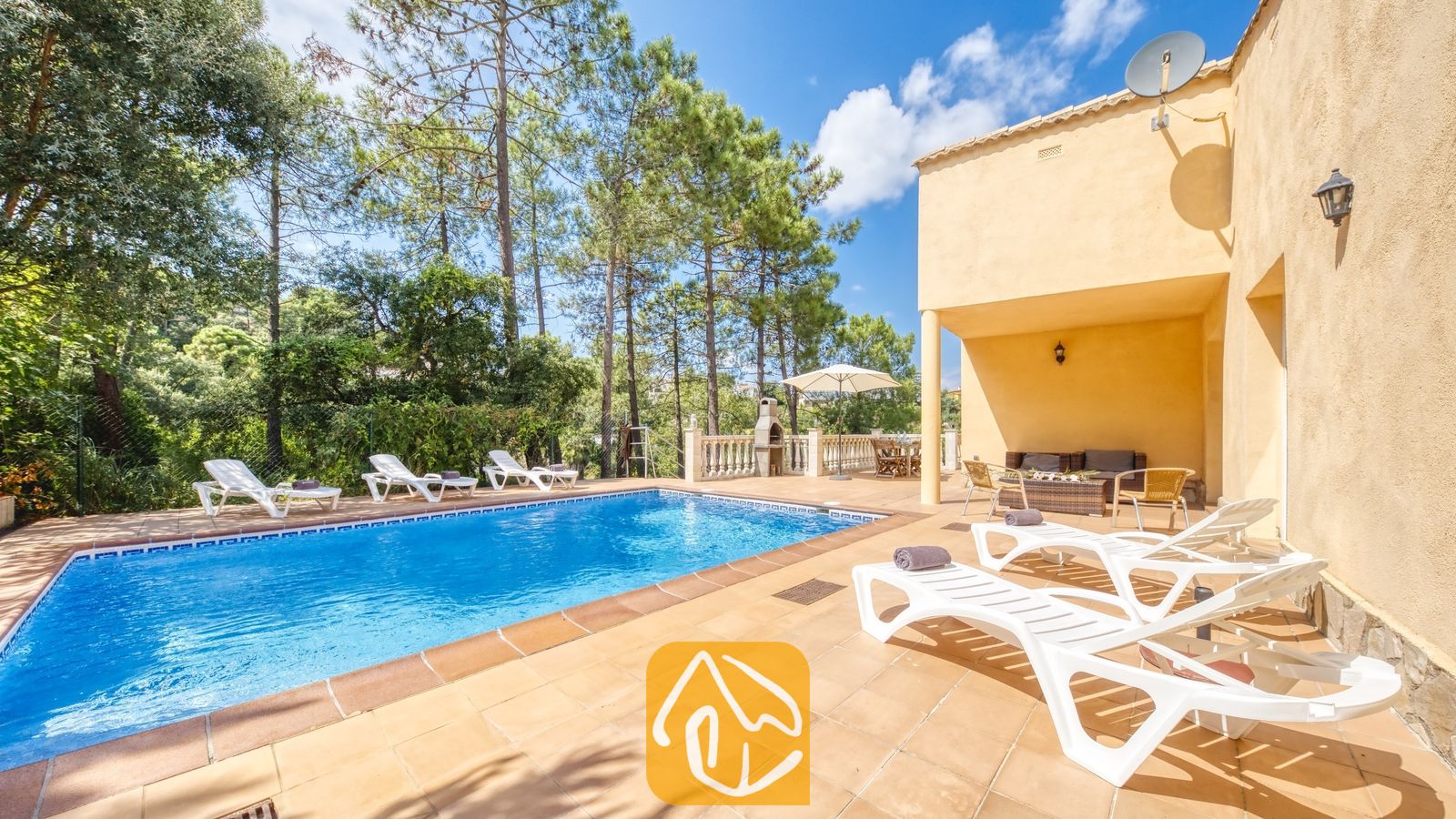 Vakantievilla's Costa Brava Spanje - Villa Esmee - Privé-buitenzwembad bij Villa Esmee met witte ligstoelen op een betegeld terras, omringd door dennenbomen onder een blauwe hemel.