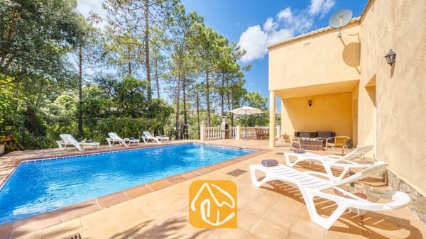 Vakantievilla's Costa Brava Spanje - Villa Esmee - Privé-buitenzwembad bij Villa Esmee met witte ligstoelen op een betegeld terras, omringd door dennenbomen onder een blauwe hemel.