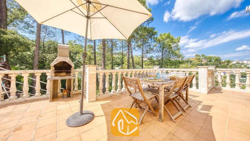 Vakantievilla's Costa Brava Spanje - Villa Esmee - Terras bij Villa Esmee, voorzien van een houten eettafel, inklapbare stoelen, een stenen barbecue en een grote parasol, met een achtergrond van dennenbomen.