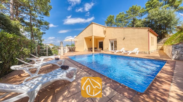 Vakantievilla's Costa Brava Spanje - Villa Esmee - Privézwembad en terras bij Villa Esmee, voorzien van witte ligstoelen met donkere handdoeken, een gele buitengevel en omringende bomen onder een blauwe hemel.