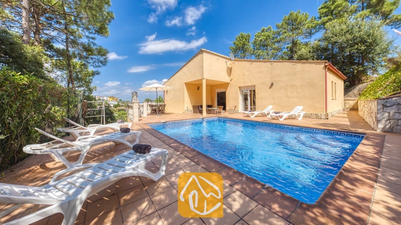 Vakantievilla's Costa Brava Spanje - Villa Esmee - Privézwembad en terras bij Villa Esmee, voorzien van witte ligstoelen met donkere handdoeken, een gele buitengevel en omringende bomen onder een blauwe hemel.