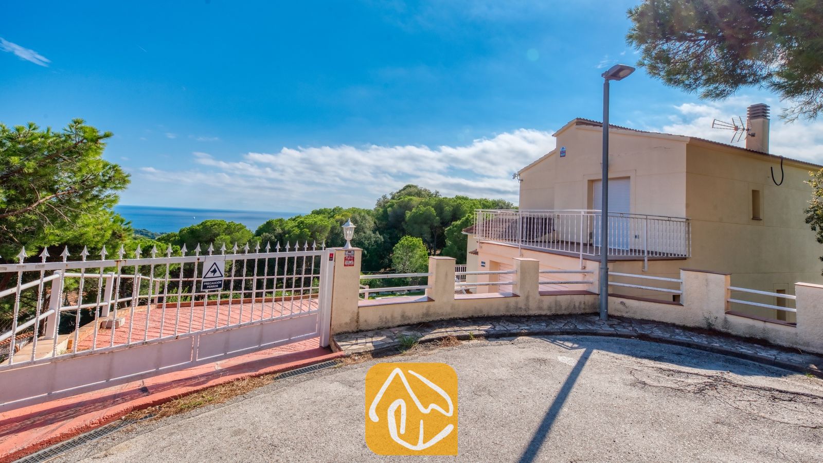 Vakantievilla's Costa Brava Spanje - Villa La Luna - Buitentoegangspoort en oprijlaan van Villa La Luna met het witte metalen hek, het gebouw met de theekleurige (of: créme/crèmekleurige) gevel en zeezicht onder een blauwe hemel.